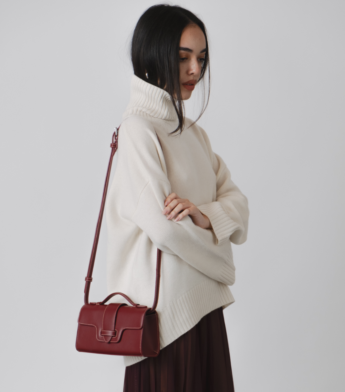 Woman wearing a cream sweater and holding a red handbag against a white background. burgundy wine handbag top handle vegan luxury bags plant based leather alternative uncaged innovations burgundy red bag WHITE RED outfit  SHEER BURGUNDY SKIRT WHITE JUMPER ROLL NECK