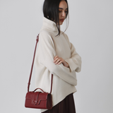 Woman wearing a cream sweater and holding a red handbag against a white background. burgundy wine handbag top handle vegan luxury bags plant based leather alternative uncaged innovations burgundy red bag WHITE RED outfit  SHEER BURGUNDY SKIRT WHITE JUMPER ROLL NECK
