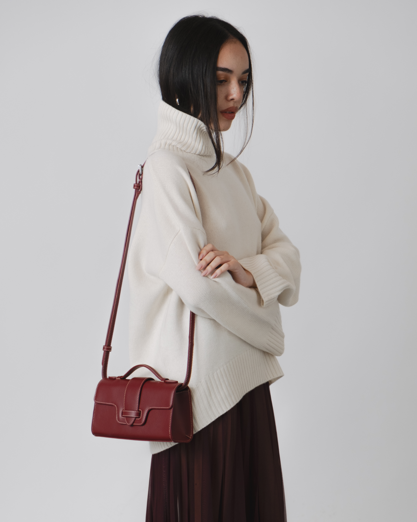 Woman wearing a cream sweater and holding a red handbag against a plain background. burgundy wine handbag top handle vegan luxury bags plant based leather alternative uncaged innovations burgundy red bag WHITE RED outfit  SHEER BURGUNDY SKIRT WHITE JUMPER ROLL NECK
