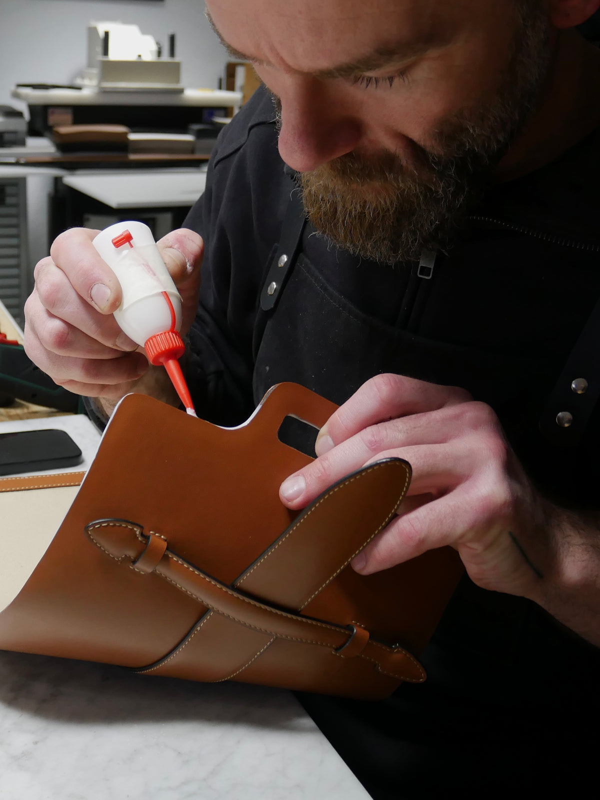 A close-up of a leather craftsman applying adhesive glue to a brown leather folio or notebook cover in a workshop setting.
